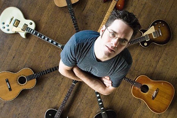 David Hillman standing, surrounded by acoustic and electric guitars in circle on floor